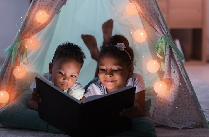 A boy and a girl under a tent lit up lamp garland are reading a book at a slumber party at Miami Children's Museum.