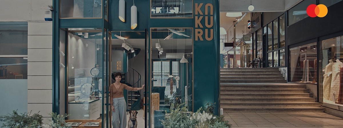 Elina Kakourou with a dog is standing at the entrance of the Kakuru jewelry boutique in Athens. There’s a Mastercard logo at the top right corner.