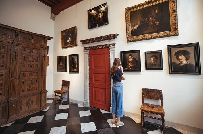 Girl looking at one of the 8 Rembrandt paintings in the Rembrandt House Museum. There is a door with 2 chairs on the sides and a wooden wardrobe in the room.