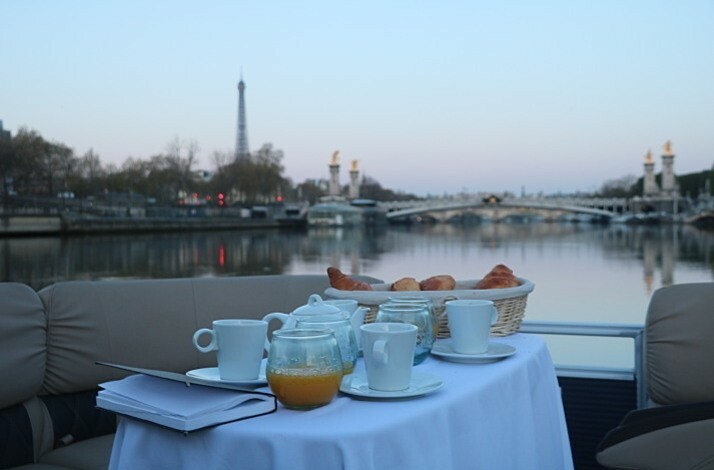 View from aboard cruise boat overlooking Seine and Eiffel Tower. On boat there is table with teapot, tea cups and croissants prepared for breakfast. 