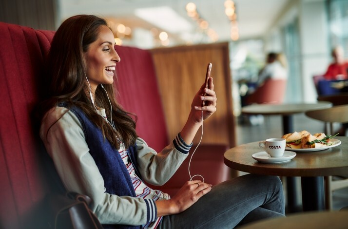 Girl is looking at her phone and laughing in the Costa Coffee cafe with a cup of espresso and sandwiches.